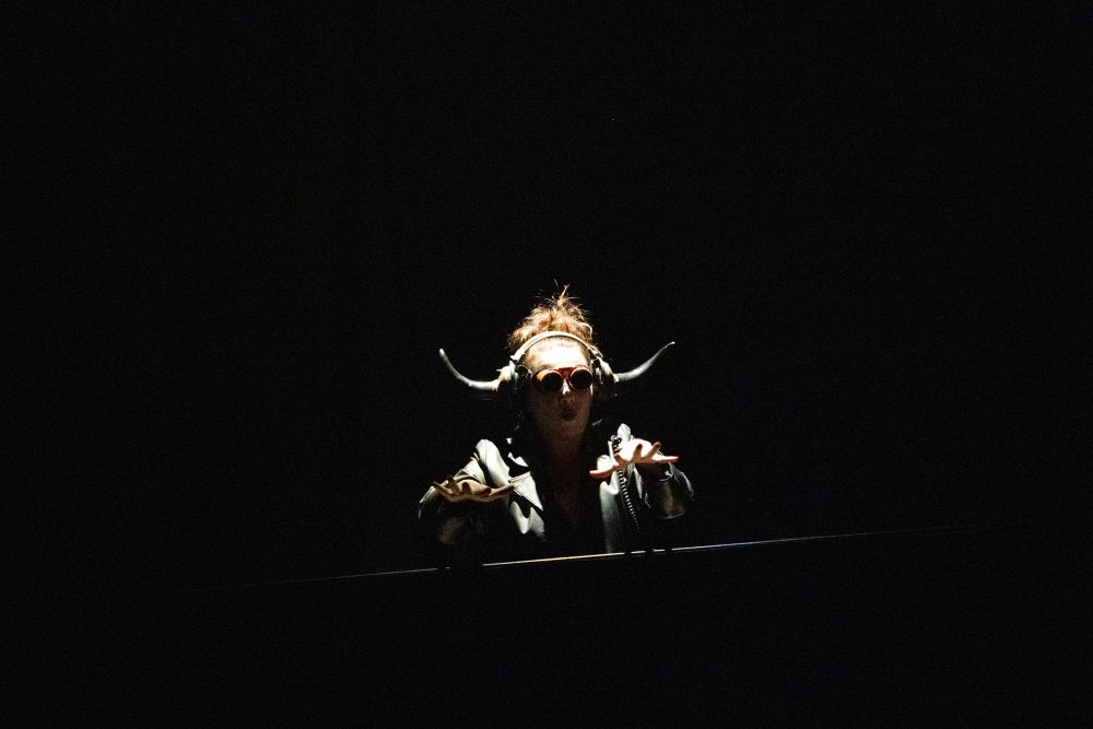 Osoba w rogatym nakryciu głowy i okrągłych okularach przeciwsłonecznych jest dramatycznie oświetlona w ciemnej przestrzeni, z podniesionymi rękami i rozłożonymi palcami, przywołując tajemniczą atmosferę spektaklu w Teatrze Witkacego.