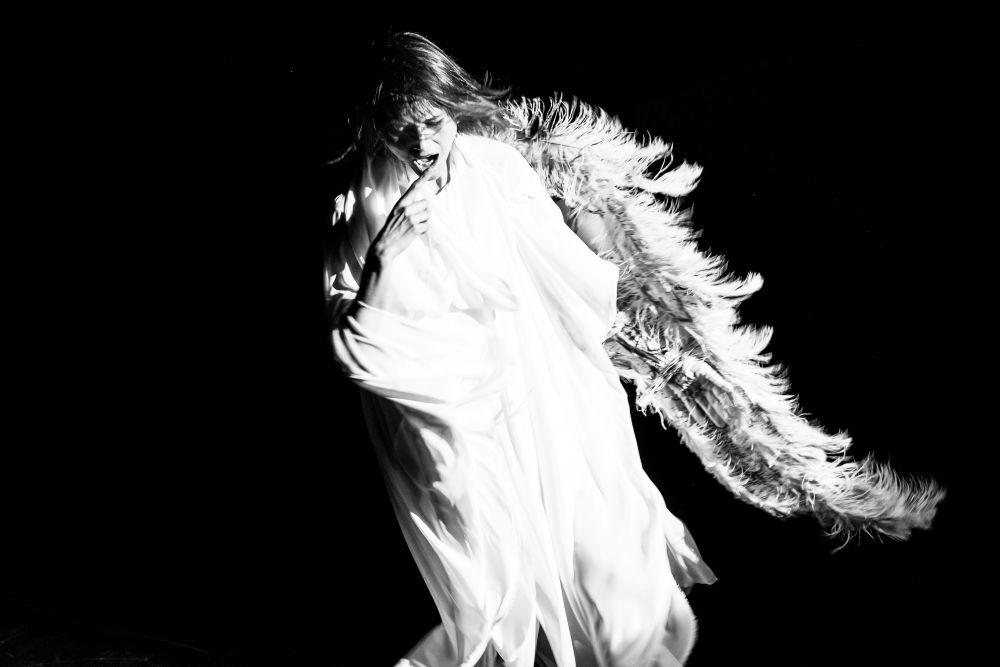 Osoba ubrana w lejącą się białą tkaninę stoi na ciemnym tle, dramatycznie oświetlona. Nawiązując do Teatru Witkacego, nosi duże pierzaste skrzydła i pozuje z ręką przy twarzy, tworząc tajemniczy, anielski wygląd.
