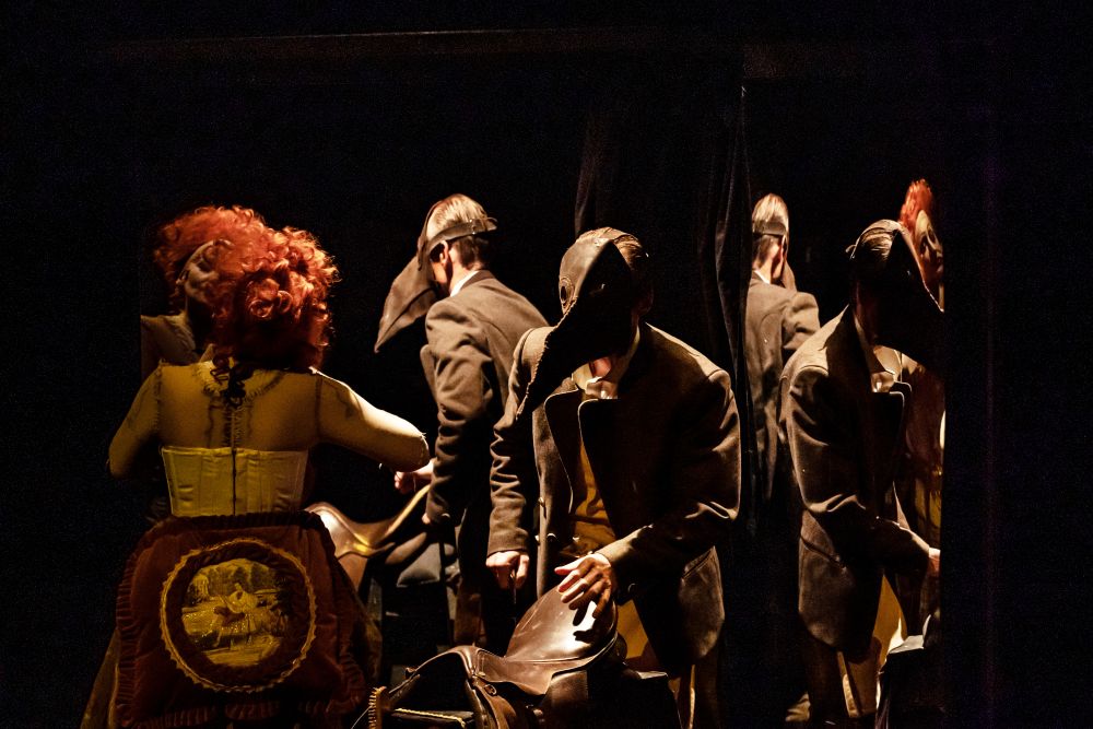 W słabo oświetlonym pokoju z lustrami zebrało się kilka osób w strojach vintage i maskach lekarzy z długimi dziobami, tworząc odbicia i surrealistyczną, teatralną atmosferę przypominającą scenę z "Metafizyki dwugłowego nieba" Teatru Witkacego.