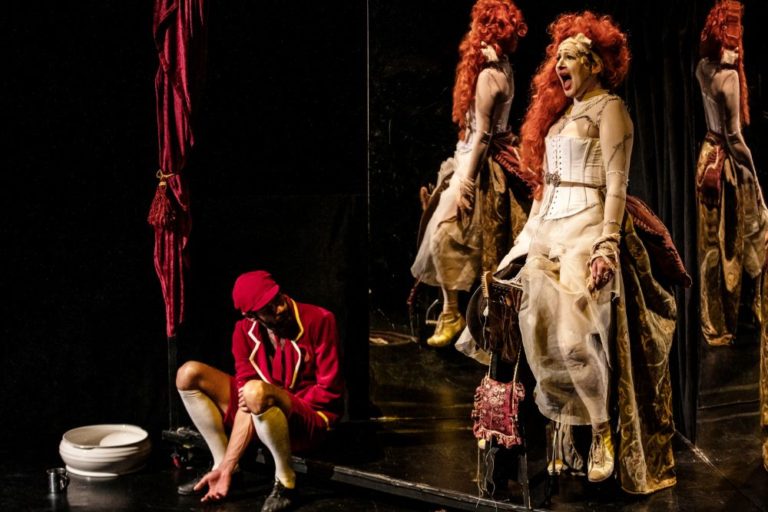 Kobieta w wyszukanym kostiumie i czerwonej peruce siedzi dramatycznie przed lustrem na scenie, podczas gdy inna w czerwonym stroju teatralnym zakłada buty obok. Ten sugestywny moment rozgrywa się w Teatrze Witkacego, oddając istotę Metafizyki dwugłowego nieba.