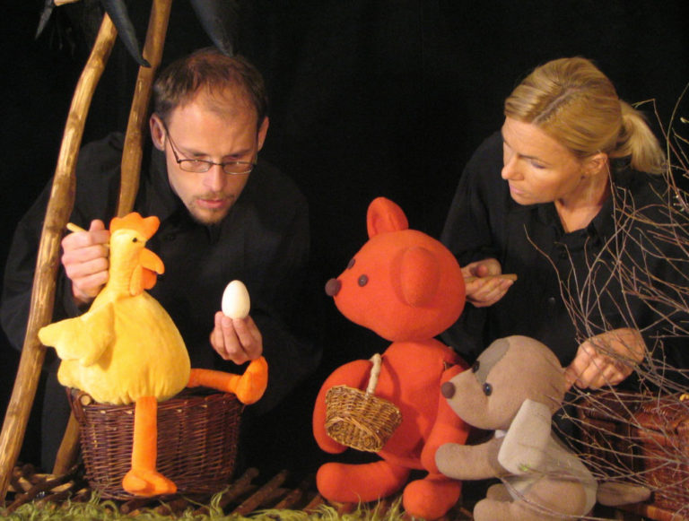 Dwóch lalkarzy ubranych na czarno ożywia żółtego kurczaka z jajkiem, czerwonego misia z koszykiem i szarą myszkę na scenie z trawy i gołych gałęzi w tym magicznym spektaklu dla fanów "Mojego Przyjaciela Pieska".