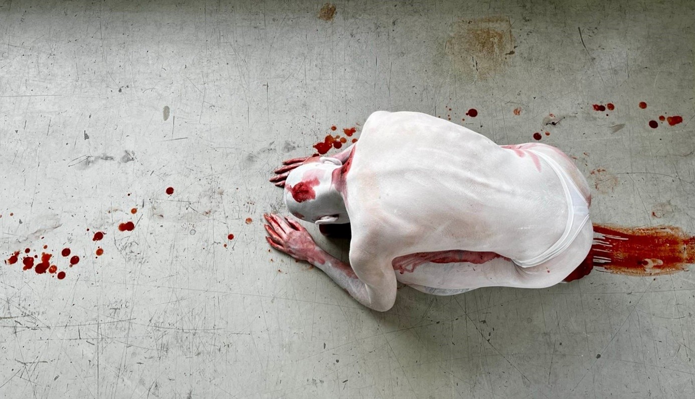Osoba pokryta białą farbą i plamami przypominającymi krew przykuca na szarej podłodze, pozostawiając za sobą rozmazany czerwony ślad i krople, przywołując obrazy przypominające felieton Pawła Głowackiego lub niepokojące gdakanie kwoki.