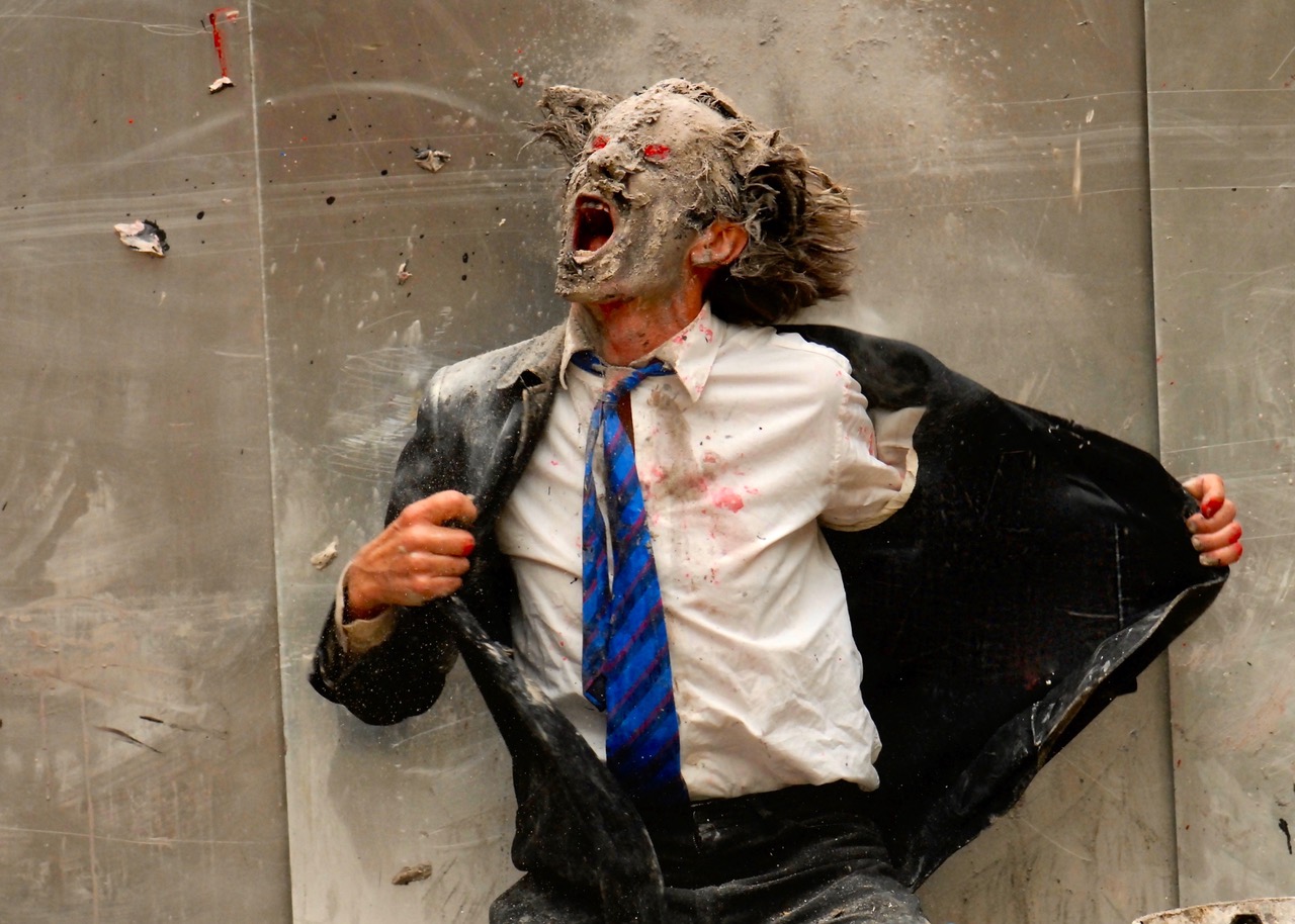 Mężczyzna w garniturze i niebieskim krawacie w paski, pokryty kurzem i gruzem, krzyczy z szeroko otwartymi ustami. Jego dzikie włosy i przygnębiony wyraz twarzy przywołują na myśl surową energię performansu na Międzyuczelnianym Przeglądzie Performansu AKTOMAR.