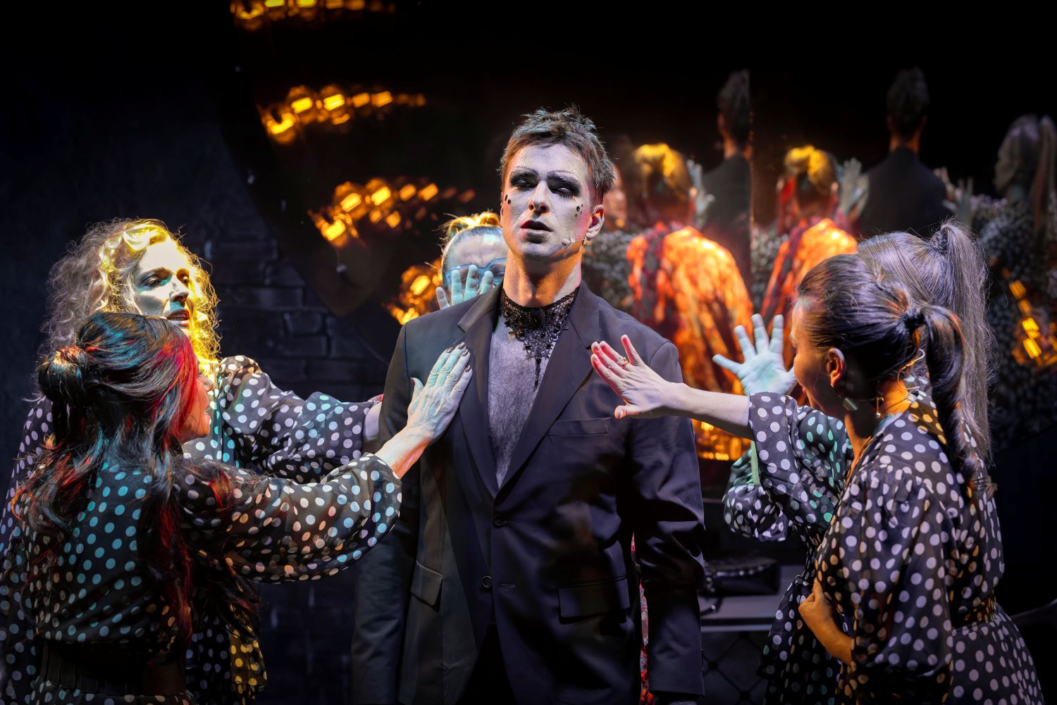 Mężczyzna w czarnym garniturze z dramatycznym makijażem stoi na środku sceny, patrząc przed siebie, a trzech wykonawców z Teatru KTO w strojach w kropki z pomalowanymi twarzami otacza go, wyciągając w jego stronę wyciągnięte ręce. Tło jest ciemne i teatralne.