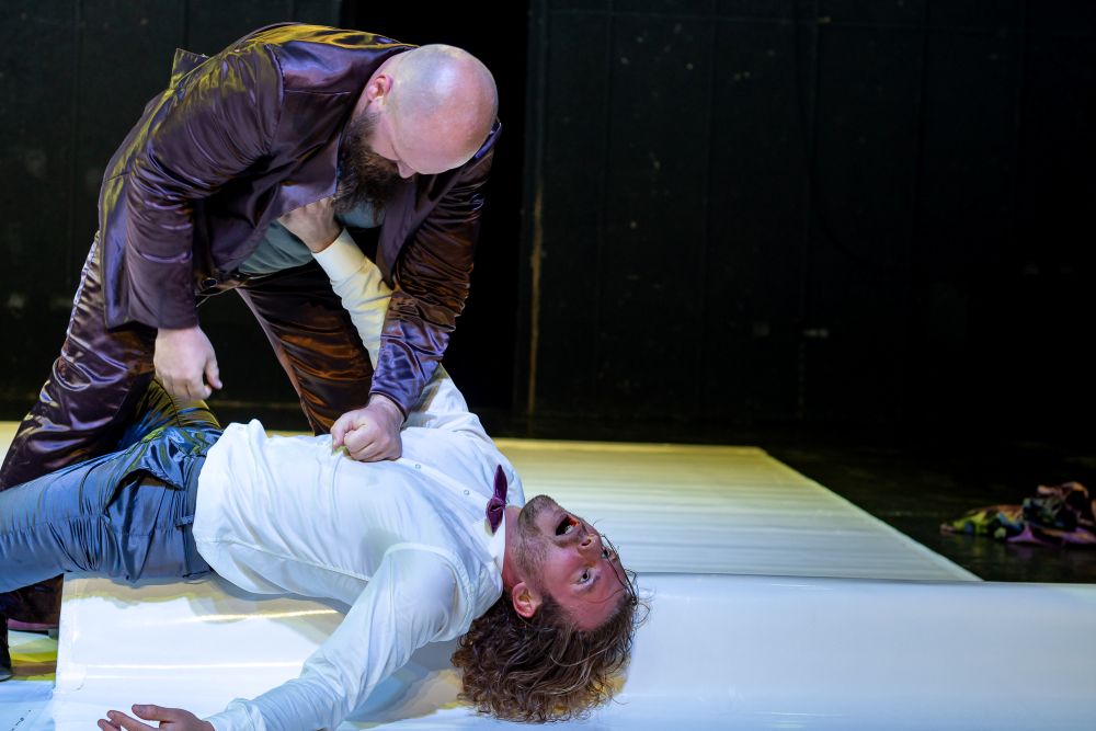 Łysy mężczyzna w błyszczącym garniturze przytrzymuje mężczyznę z kręconymi włosami i w białej koszuli, który leży na podłodze, patrząc w górę w szoku lub strachu, podczas trzymającej w napięciu sceny na słabo oświetlonej scenie Teatru KTO.