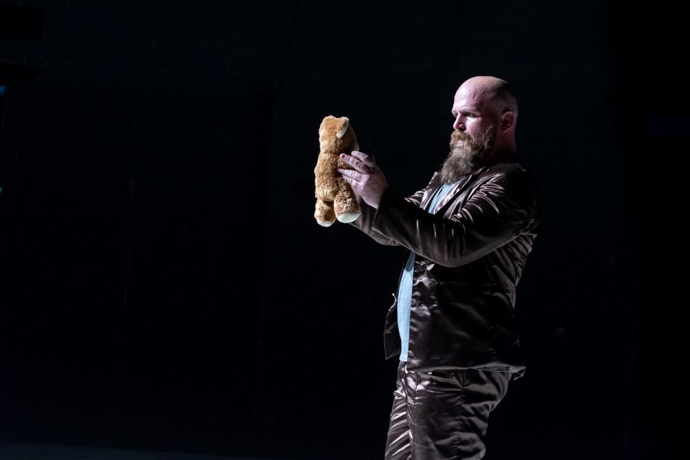 Brodaty mężczyzna w brązowym aksamitnym garniturze stoi na ciemnej scenie w Teatrze KTO, trzymając małego brązowego misia obiema rękami i patrząc na niego uważnie.