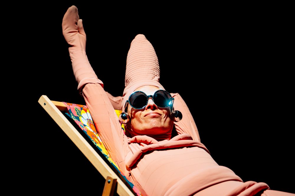 Osoba ubrana w różowy kostium w kształcie stożka i duże okrągłe okulary przeciwsłoneczne leży na kolorowym leżaku na czarnym tle, z rękami uniesionymi figlarnie nad głową.