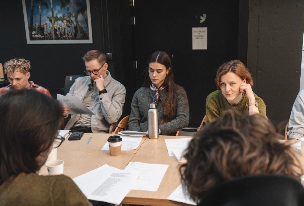 Grupa ludzi siedzi wokół stołu z papierami, filiżankami kawy i butelką wody, wyglądając na skupionych i zaangażowanych w spotkanie dotyczące premiery ruchu Plagi w nowoczesnym pomieszczeniu.