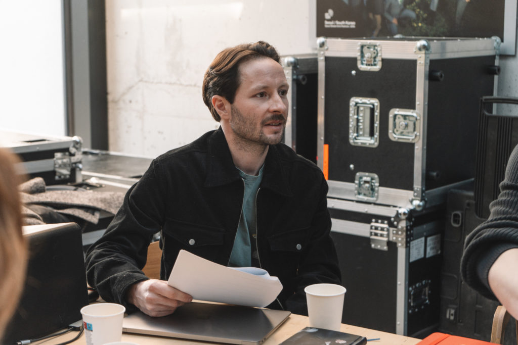 Mężczyzna z krótkimi brązowymi włosami i przystrzyżoną brodą siedzi przy stole i trzyma dokumenty, być może przeglądając próby przed zbliżającą się premierą. Ma na sobie czarną kurtkę i zieloną koszulę. W tle widać dwa papierowe kubki, telefon i obudowy urządzeń elektronicznych.