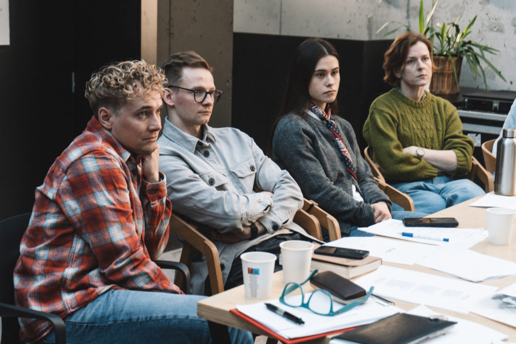 Cztery osoby siedzą przy stole z papierami, notatnikami i kubkami, skupione na czymś, co wygląda na poważne spotkanie. Otoczenie sugeruje nowoczesny warsztat - być może omawianie prób lub finalizowanie planów na nadchodzącą premierę Plagi.