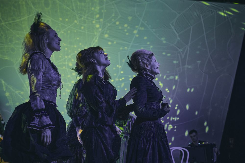 Trzy kobiety w ciemnych kostiumach w stylu vintage śpiewają na scenie z zielonymi, świecącymi wzorami świetlnymi za nimi podczas koncertu inspirowanego Bertoltem Brechtem. W tle widać grającego muzyka.