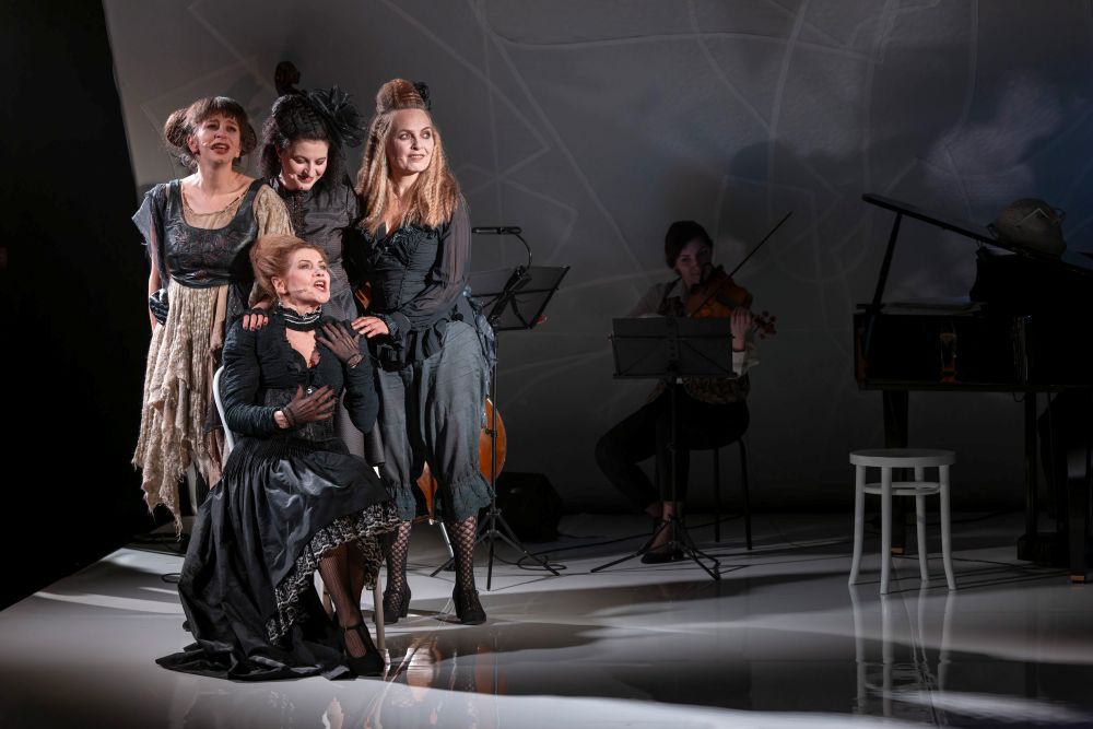 Cztery kobiety w teatralnych, czarnych kostiumach w stylu vintage występują dramatycznie na scenie, przywołując klimat Bertolta Brechta, podczas gdy dwóch muzyków w tle gra na instrumentach smyczkowych pośród dramatycznego oświetlenia i wzorów cieni na ścianie.