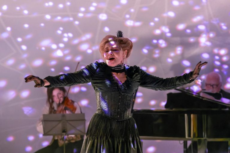 Kobieta w czarnej sukience śpiewa namiętnie na scenie z wyciągniętymi ramionami podczas koncertu. Za nią grają skrzypek i pianista, a fioletowe i białe wzory świetlne tworzą dramatyczny efekt przywołujący teatralny styl Bertolta Brechta.