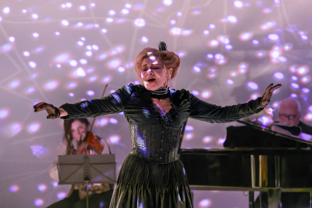 Kobieta w czarnej sukience śpiewa namiętnie na scenie z wyciągniętymi ramionami podczas koncertu. Za nią grają skrzypek i pianista, a fioletowe i białe wzory świetlne tworzą dramatyczny efekt przywołujący teatralny styl Bertolta Brechta.