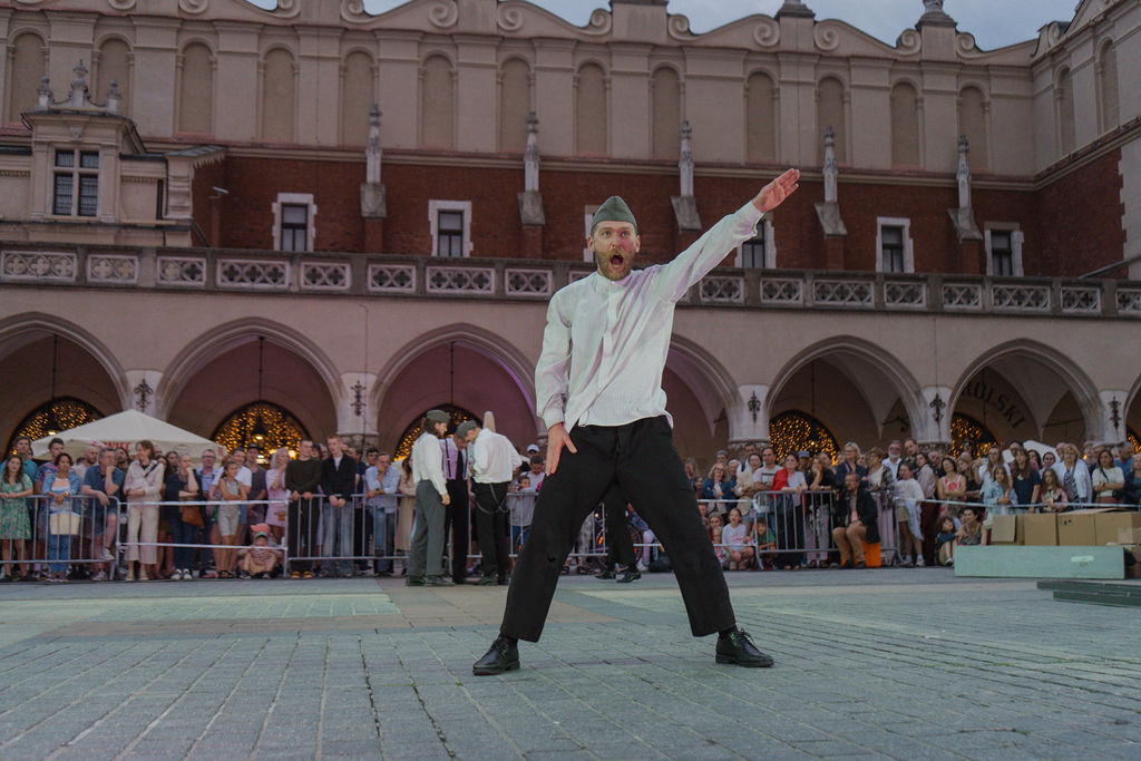 Uliczny performer w białej koszuli i ciemnych spodniach, część międzynarodowej trasy teatralnej KTO ON TOUR 2026, tańczy z podniesioną ręką i otwartymi ustami przed dużym tłumem na historycznym placu miejskim z łukowatymi budynkami za plecami.