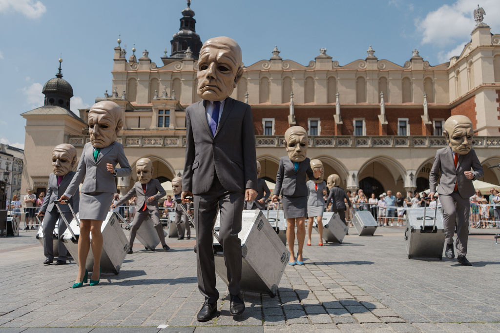 Uliczni performerzy z zespołu teatralnego, będącego częścią międzynarodowej trasy teatralnej KTO ON TOUR 2026, chodzą w garniturach i zbyt dużych maskach, a każdy z nich ciągnie metalową walizkę na miejskim placu z zabytkowym budynkiem i błękitnym niebem za plecami.