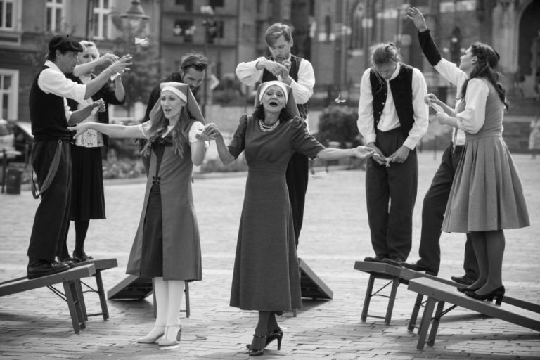 Grupa performerów z Teatru KTO w strojach w stylu vintage zachowuje się i śpiewa ekspresyjnie na ławkach na świeżym powietrzu na czarno-białym zdjęciu z Unterwegs Theaterfestival, z dwiema kobietami z przodu i pośrodku, podczas gdy inni gestykulują dramatycznie za nimi.
