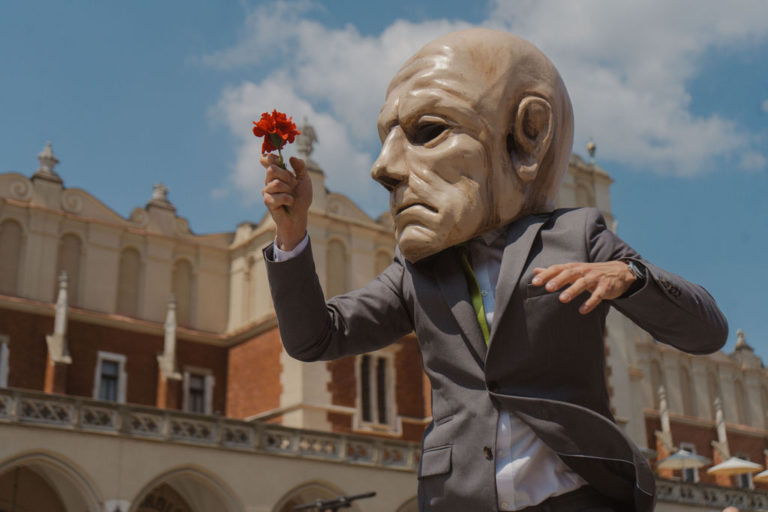 Performer w garniturze nosi dużą, przerysowaną maskę na głowę i trzyma czerwone kwiaty, pozując dramatycznie w plenerze przed ozdobnym budynkiem pod błękitnym niebem - oddając ducha teatru na międzynarodowej trasie teatralnej KTO ON TOUR 2026.