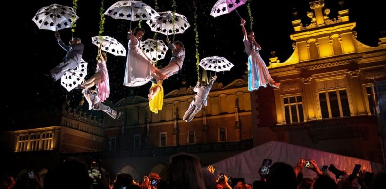 Performerzy w białych kostiumach z Teatru KTO zwisają z otwartymi parasolami nad nocnym tłumem. Oświetlony zabytkowy budynek wzmacnia intensywność sceny, gdy widzowie oglądają i rejestrują pokaz, podziwiając teatralną odwagę.