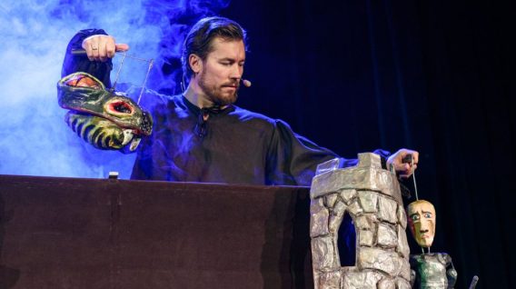 Lalkarz w ciemnym ubraniu wykonuje BAŚŃ O RYCERZU BEZ KONIA, zręcznie operując lalką smoka i lalką rycerza w pobliżu kamiennej wieży na scenie, z niebieskim dymem wirującym w tle.