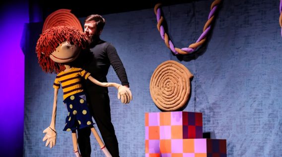 Lalkarz w czerni manipuluje dużą lalką - zmyślonym chłopcem z włosami z czerwonej włóczki, żółtą koszulą w paski i niebieską spódnicą w kropki. Kolorowe rekwizyty i niebieska kurtyna ze skręconym sznurem tworzą scenę przypominającą baśń.