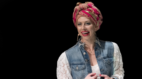 Kobieta uśmiecha się podczas rozmowy. Nosi żywą różową opaskę na głowę, duże kolczyki w kształcie kół, dżinsową kamizelkę na białej koronkowej koszuli i ma czerwoną szminkę i paznokcie. Tło jest czarne.