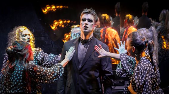 Mężczyzna w czarnym garniturze z dramatycznym makijażem stoi na środku sceny, patrząc przed siebie, a trzech wykonawców z Teatru KTO w strojach w kropki z pomalowanymi twarzami otacza go, wyciągając w jego stronę wyciągnięte ręce. Tło jest ciemne i teatralne.