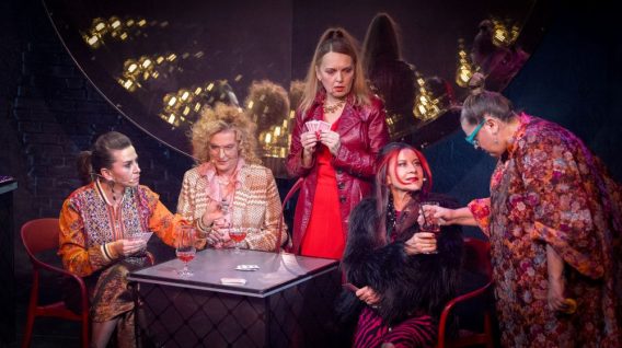 Pięć osób siedzi przy małym stoliku i gra w karty, ubranych w kolorowe, krzykliwe stroje. Jedna osoba stoi i wydaje się rozmawiać z pozostałymi. Scena rozgrywa się w słabo oświetlonym, dramatycznym otoczeniu. Pięć osób siedzi przy małym stoliku i gra w karty, ubranych w kolorowe, krzykliwe stroje. Jedna osoba stoi i wydaje się rozmawiać z pozostałymi. Scena rozgrywa się w słabo oświetlonym, dramatycznym otoczeniu.