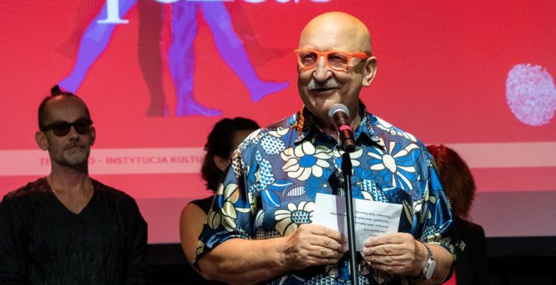 Łysy mężczyzna z wąsami, w czerwonych okularach i kwiecistej koszuli, przemawia do mikrofonu na scenie. Za nim stoją trzy osoby. W tle wyświetlany jest polski tekst na czerwonym ekranie.