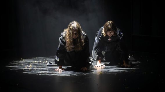 Dwie osoby w ciemnych, dramatycznych strojach kucają na słabo oświetlonej scenie Teatru KTO, z rękami na ziemi i opuszczonymi głowami. Reflektor tworzy cienie i podkreśla mglistą, nastrojową scenerię.