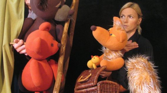 Kobieta występuje w spektaklu "Mój Przyjaciel Pies" z trzema dużymi lalkami zwierząt - niedźwiedziem brunatnym, niedźwiedziem pomarańczowym i pomarańczowym lisem z puszystym ogonem - obok kosza i kurtyny, na czarnym tle.