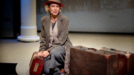 Kobieta w ubraniu vintage i czerwonym kapeluszu klęczy na scenie obok dwóch starych walizek, patrząc nieco poza kamerę, z neutralnym tłem i dużą białą kolumną za nią. Kobieta w ubraniu vintage i czerwonym kapeluszu klęczy na scenie obok dwóch starych walizek, patrząc nieco poza kamerę, z neutralnym tłem i dużą białą kolumną za nią.