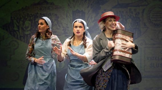 Trzy kobiety na scenie, dwie w niebieskich fartuchach i chustach na głowach, wyglądające na zaniepokojone, a jedna w kapeluszu i płaszczu uśmiechająca się, trzymając duży cylindryczny pakiet, z żółto oświetlonym tłem za nimi.