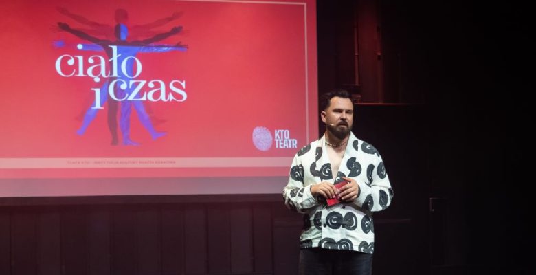 Na scenie stoi brodaty mężczyzna trzymający czerwony przedmiot, przemawiający przed dużym ekranem z napisem ciało i czas, abstrakcyjnymi ludzkimi sylwetkami i logo KTO Teatr wyświetlanymi na żywym czerwonym tle.
