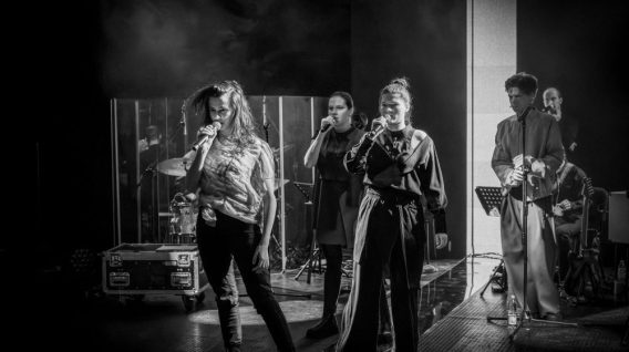 Czarno-białe zdjęcie pięciu muzyków występujących na scenie. Trzech śpiewaków stoi z przodu z mikrofonami, podczas gdy dwóch muzyków gra na instrumentach w tle, przywołując ponadczasowy dramat sonety szekspira wśród świateł i wirującego dymu.