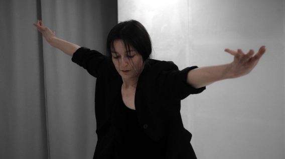Osoba ubrana na czarno z ciemnymi włosami stoi na jasnym tle, z szeroko rozłożonymi ramionami i lekko pochyloną głową, badając ciało i czas w ekspresyjnej pozie tanecznej.