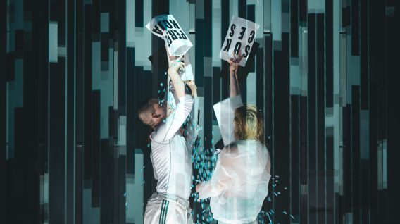 Dwie osoby w białych ubraniach stoją przed pasiastym tłem, trzymając i wyrywając białe znaki z czarnymi literami - scena z filmu Spektakle: Teoria Zmęczenia. Niebieskie konfetti rozsypuje się w powietrzu wokół nich.