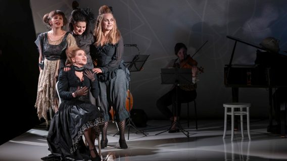 Cztery kobiety w dramatycznych czarno-szarych sukniach występują na scenie; trzy stoją, a jedna klęczy. Za nimi muzyk podnosi instrument smyczkowy obok fortepianu i białego stołka przy słabym oświetleniu, tworząc sugestywną scenę ze współczesnego spektaklu.