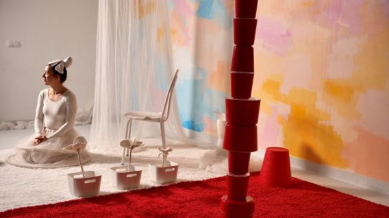 Kobieta w białym stroju i chustce na głowie siedzi na białym dywanie, patrząc w lewo. Za nią znajduje się białe krzesło, przezroczysta tkanina i pastelowa fototapeta. Na pierwszym planie na czerwonym dywanie ułożone są czerwone wiadra, a obok nich białe wiadra i sztuczne kwiaty.