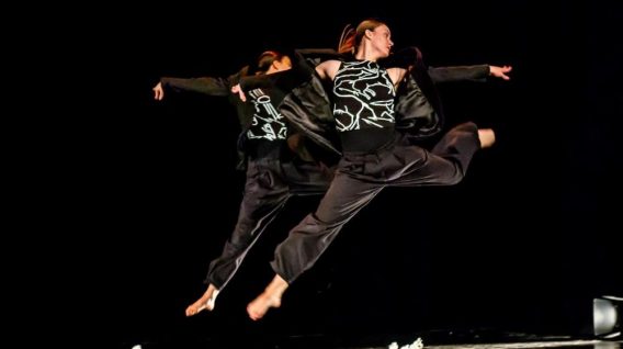 Dwóch tancerzy w czarnych strojach energicznie skacze po ciemnej scenie, a ich ekspresyjne ruchy zostały uchwycone w powietrzu. Tło jest czarne, co podkreśla, jak ciało i czas łączą się w ich dynamicznych pozach.