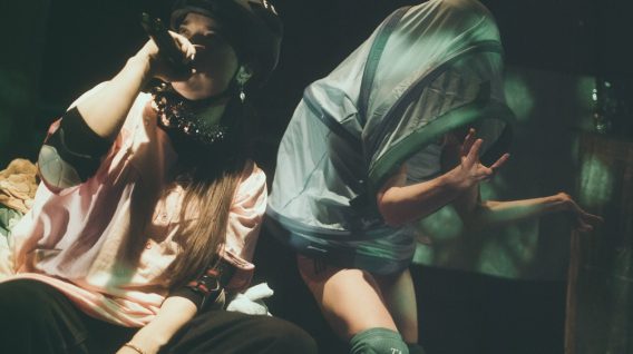 Dwóch wykonawców na scenie: jeden siedzi z mikrofonem w kasku i różowej koszuli, podczas gdy drugi, w szortach i nakolannikach, dramatycznie pozuje pod prześwitującym materiałem – uderzający moment z wyjątkowego pokazu Spektakle. Dwóch wykonawców na scenie: jeden siedzi z mikrofonem w kasku i różowej koszuli, podczas gdy drugi, w szortach i nakolannikach, dramatycznie pozuje pod prześwitującym materiałem - uderzający moment z wyjątkowego pokazu Spektakle.