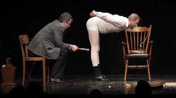 Dwóch mężczyzn występuje na scenie. Jeden mężczyzna w garniturze siedzi i trzyma gazetę w pobliżu drugiego mężczyzny, który jest pochylony w białej bieliźnie i skarpetkach. Widoczne są krzesła, porozrzucane śmieci i rozbity wazon.