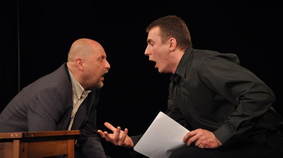 Dwóch mężczyzn w ciemnych ubraniach siedzi przy stole, zwróceni do siebie z intensywnymi wyrazami twarzy, z otwartymi ustami, jakby kłócili się lub namiętnie dyskutowali. Jeden z nich trzyma kartkę papieru. Tło jest czarne.