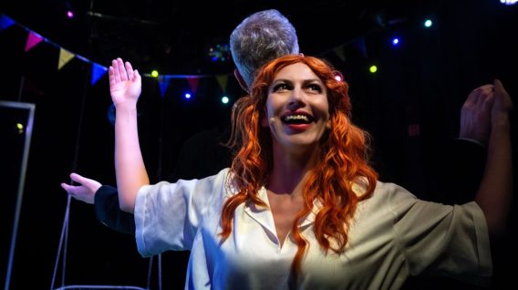 Uśmiechnięta kobieta z długimi rudymi włosami i białą koszulą unosi ręce, stojąc przed mężczyzną z siwymi włosami pod kolorowymi lampkami i świątecznymi chorągiewkami w ciemnym otoczeniu.